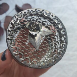 Puede incluir: Una caja de metal plateado con un dise&ntilde;o floral decorativo. La caja tiene una parte superior de malla y un borde festoneado.