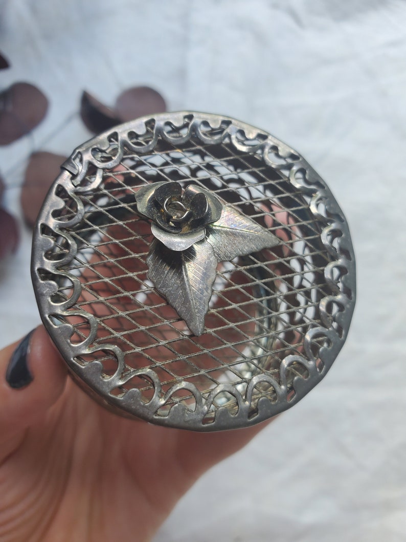 Puede incluir: Una caja de metal plateado con un dise&ntilde;o floral decorativo. La caja tiene una parte superior de malla y un borde festoneado.