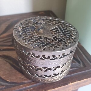 Puede incluir: Una caja de metal plateado con un dise&ntilde;o floral decorativo y una tapa de malla. La caja es redonda y tiene un dise&ntilde;o delicado e intrincado.