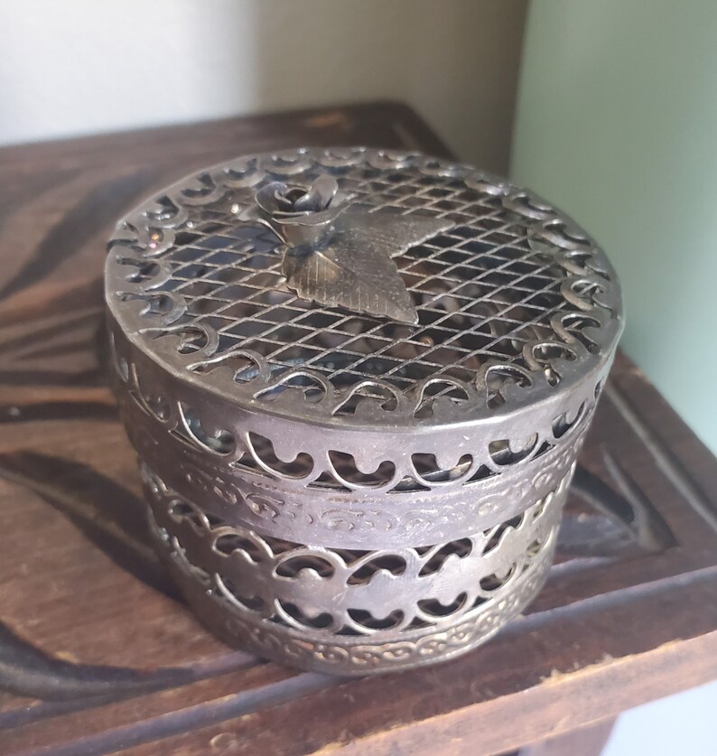 Puede incluir: Una caja de metal plateado con un dise&ntilde;o floral decorativo y una tapa de malla. La caja es redonda y tiene un dise&ntilde;o delicado e intrincado.