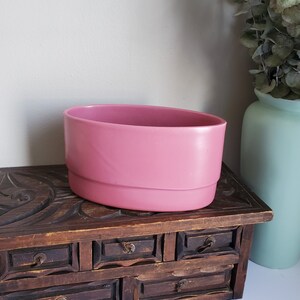 May include: A pink oval ceramic planter with a slightly raised lip around the top edge. The planter is sitting on a wooden chest with carved details and multiple drawers.