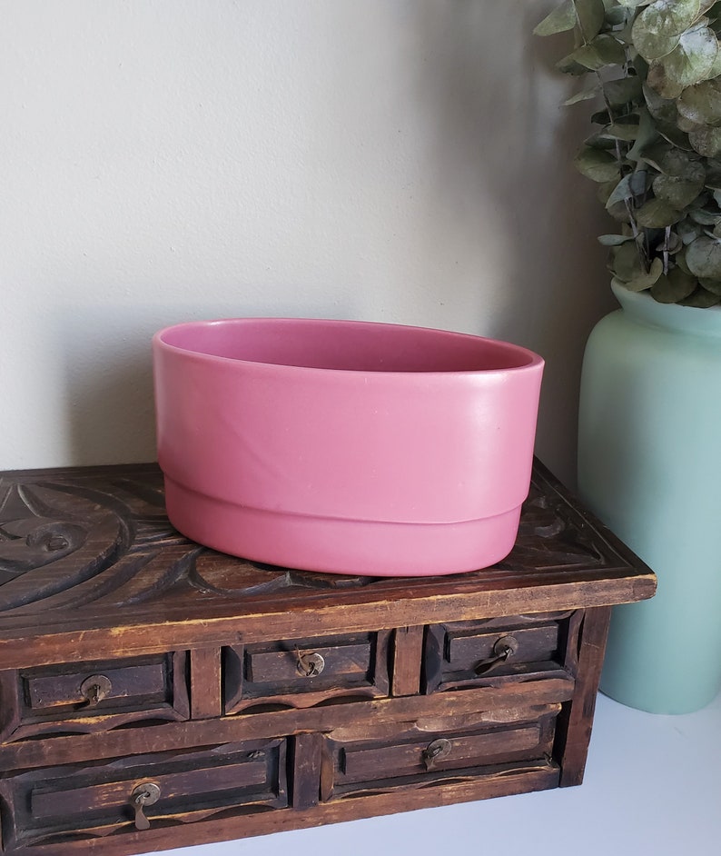 May include: A pink oval ceramic planter with a slightly raised lip around the top edge. The planter is sitting on a wooden chest with carved details and multiple drawers.
