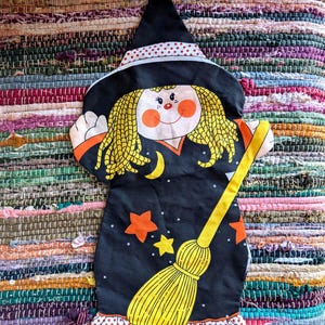 May include: A flat, fabric witch decoration featuring a black pointed hat with a red and white polka dot band, yellow yarn-style hair, and a smiling face. The black dress displays orange and yellow stars, a yellow crescent moon, and a broom.