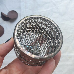 Puede incluir: Un porta anillos de metal plateado con un dise&ntilde;o decorativo, intrincado y geom&eacute;trico. El porta anillos est&aacute; hecho de una fina malla de alambre y tiene una forma circular.