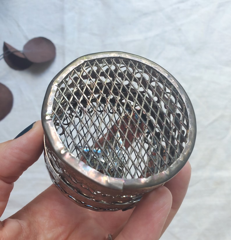 Puede incluir: Un porta anillos de metal plateado con un dise&ntilde;o decorativo, intrincado y geom&eacute;trico. El porta anillos est&aacute; hecho de una fina malla de alambre y tiene una forma circular.