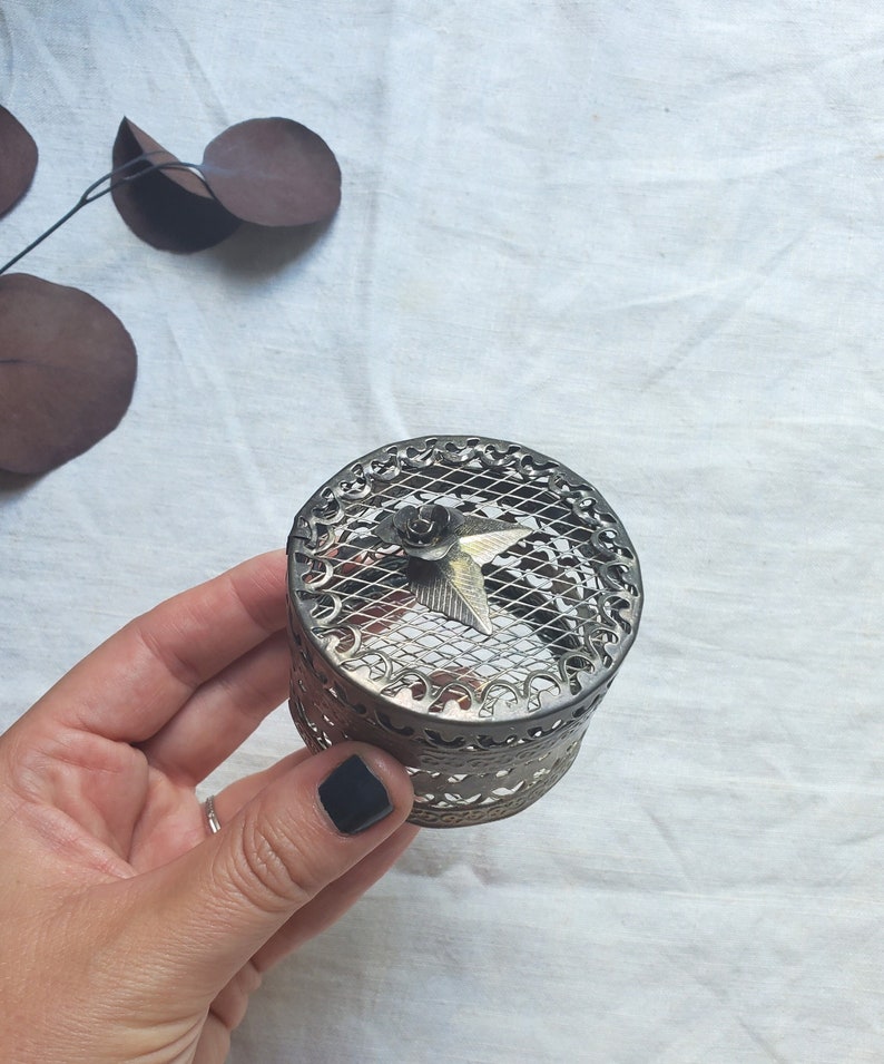 Puede incluir: Una caja de metal plateado para guardar peque&ntilde;os objetos con un dise&ntilde;o decorativo e intrincado. La tapa presenta un patr&oacute;n de malla y una peque&ntilde;a flor dorada con un centro en forma de estrella.