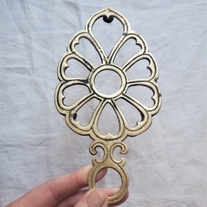May include: A gold-toned metal hot pad with a decorative floral design. The pad has a ring handle for easy carrying.