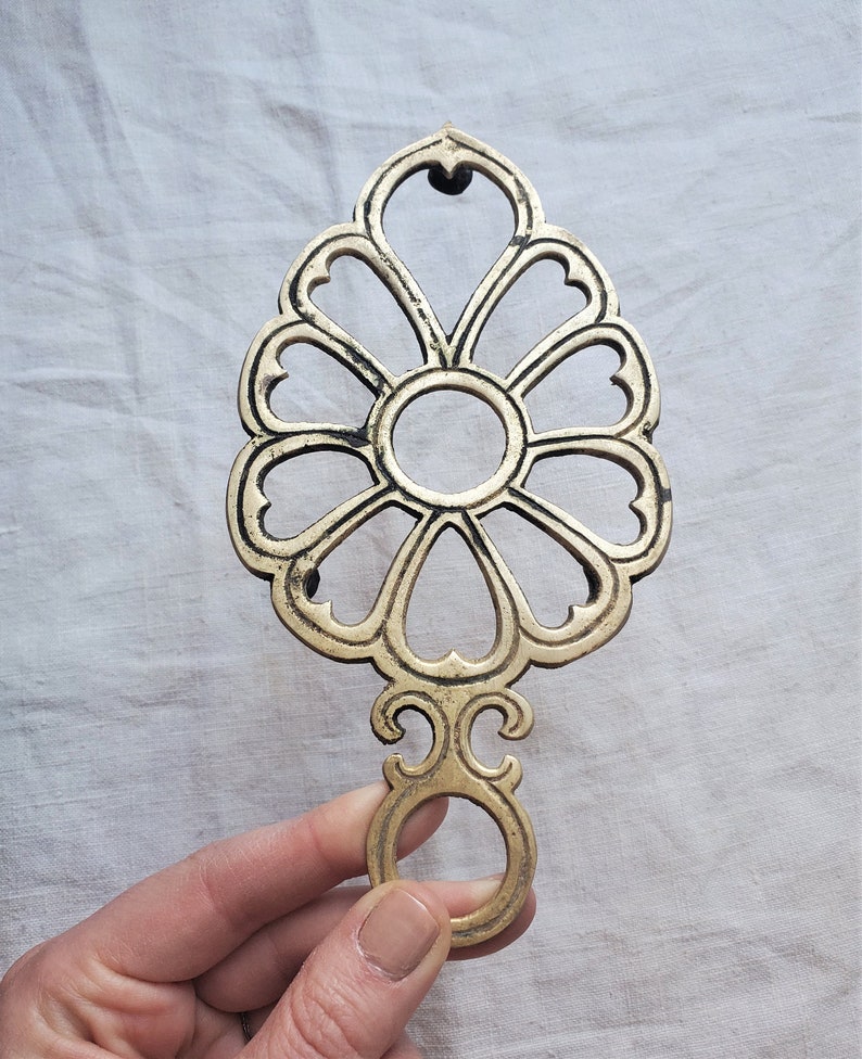 May include: A gold-toned metal hot pad with a decorative floral design. The pad has a ring handle for easy carrying.
