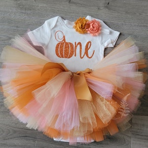 May include: A white onesie with a glittery orange pumpkin and the word "one" on it. The onesie is paired with a multi-layered tulle skirt in shades of orange, peach, and cream. The skirt has a gold satin bow.