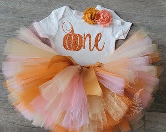May include: A white onesie with a glittery orange pumpkin and the word "one" on it. The onesie is paired with a multi-layered tulle skirt in shades of orange, peach, and cream. The skirt has a gold satin bow.