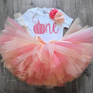 May include: A white baby bodysuit with a pink glitter pumpkin graphic and the word "one". The outfit includes a layered tutu skirt in shades of pink and peach, and is adorned with a pink flower.