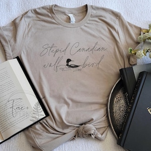 May include: Beige t-shirt with the text "Stupid Canadian wolf bird" and a small illustration of a loon. The shirt is knotted at the waist and is displayed with an open book, a decorative tray, and two hardback books.
