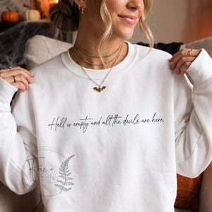 May include: A white long-sleeved crewneck sweatshirt featuring the black cursive text Hell is empty and all the devils are here printed across the chest. The garment has a simple round neckline and long sleeves, providing a clean and neutral base.