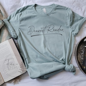 May include: A pale green t-shirt with the words "Dearest Reader," in black script. The shirt is knotted at the bottom. An open book and a decorative tray with a book are also in the image.