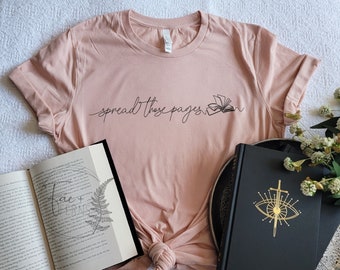 Può includere: Maglietta rosa chiaro con la scritta "spread those pages" in corsivo nero e un'illustrazione di un libro. L'immagine presenta libri aperti e un diario nero con un disegno di occhio dorato, suggerendo un tema letterario.