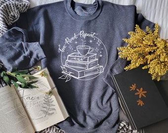 May include: A dark gray sweatshirt with the words "Tea, Read, Repeat" in a white circular design. The design includes a stack of books, a teacup, and stars. An open book and a black book are also visible.