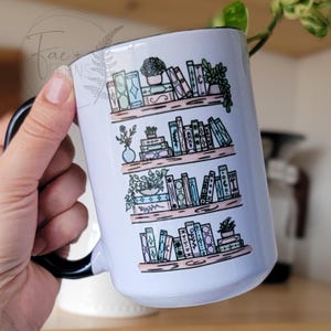 May include: White ceramic mug with a black handle featuring a colourful illustration of a bookshelf filled with books and plants. The bookshelf has four shelves and is decorated with flowers, leaves, and small pots.