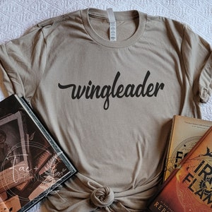 May include: A beige t-shirt with the text "wingleader" printed in black cursive font. The shirt is laid out on a white surface with two books.