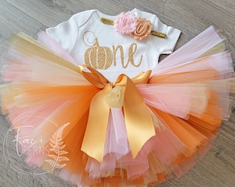 Könnte beinhalten: Ein weißer Strampler mit einem goldenen Glitzerkürbis und dem Wort "one" darauf. Der Strampler wird mit einem Tüll-Tutu in Orange-, Rosa- und Goldtönen kombiniert. Ein goldenes Stirnband mit rosa und pfirsichfarbenen Blumen vervollständigt das Outfit.