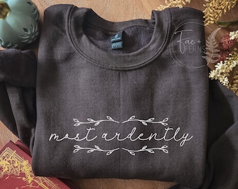 May include: A dark charcoal gray crewneck sweatshirt featuring the white text "most ardently" in a cursive script, flanked by delicate white line-drawn leafy branches. The garment has a ribbed collar and long sleeves, with a visible brand tag.