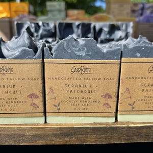 May include: Close-up of handcrafted tallow soap bars. The soap has a gray and white marbled appearance and is labeled "Geranium + Patchouli." Each bar is 4.5 oz and made with locally rendered beef suet. The brand name "Soap Roots" is visible.