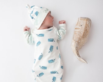 May include: A white baby swaddle with a blue whale print. The swaddle is tied at the bottom and has a matching whale-print hat. A wooden whale toy is on the right side of the image.