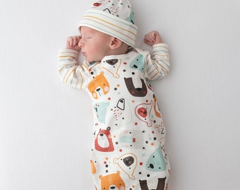 May include: A white baby sleeping bag with a colorful pattern of bears, foxes, and other woodland creatures. The sleeping bag has long sleeves and a fold-over foot. The baby is wearing a white and yellow striped hat.