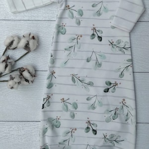 May include: A white baby gown with a botanical print. The gown features a pattern of green leaves and small brown berries, set against a backdrop of horizontal gray stripes. The sleeves are striped, and the gown is made of soft fabric.
