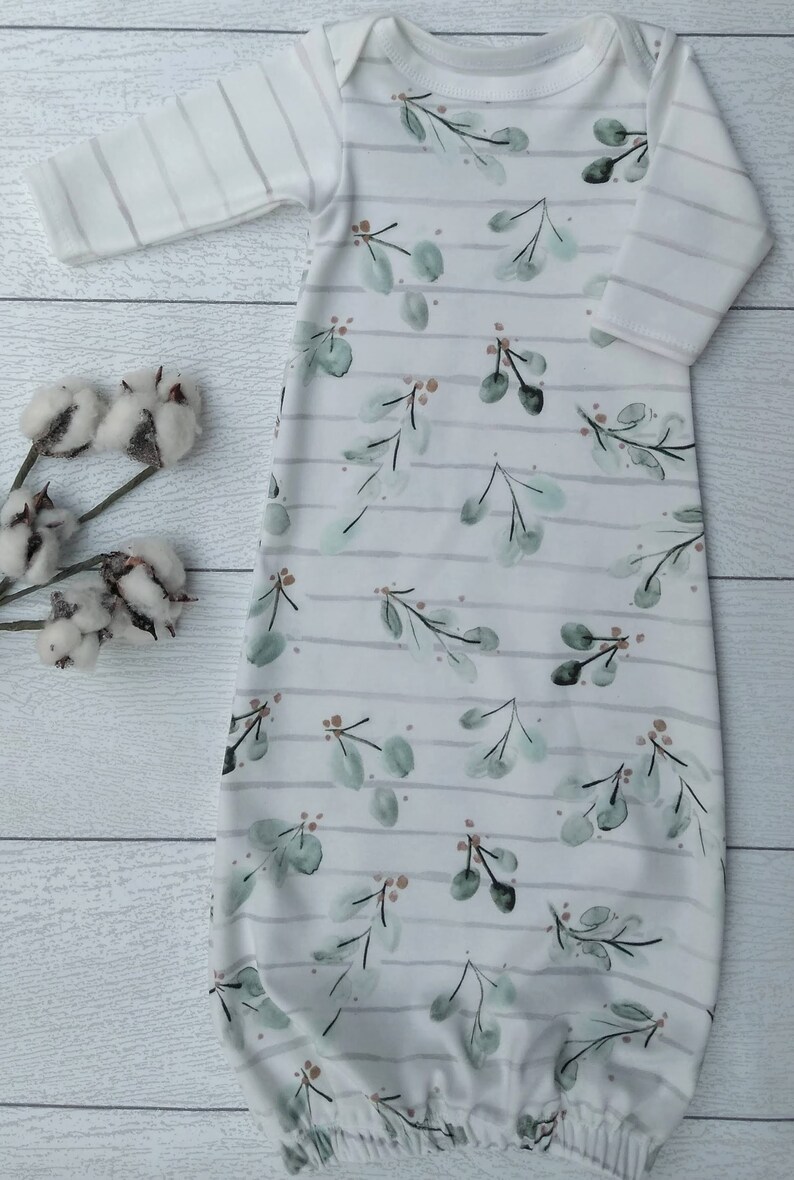 May include: A white baby gown with a botanical print. The gown features a pattern of green leaves and small brown berries, set against a backdrop of horizontal gray stripes. The sleeves are striped, and the gown is made of soft fabric.