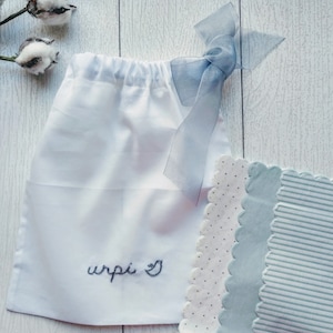 May include: A white drawstring bag with the embroidered word "wrpi" and a heart symbol. The bag is tied with a light blue ribbon. Beside the bag are three decorative fabric pieces with scalloped edges, featuring polka dots and stripes.