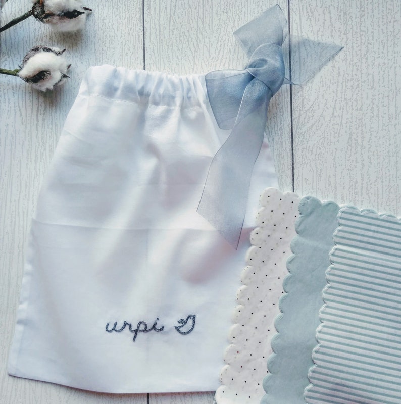 May include: A white drawstring bag with the embroidered word "wrpi" and a heart symbol. The bag is tied with a light blue ribbon. Beside the bag are three decorative fabric pieces with scalloped edges, featuring polka dots and stripes.