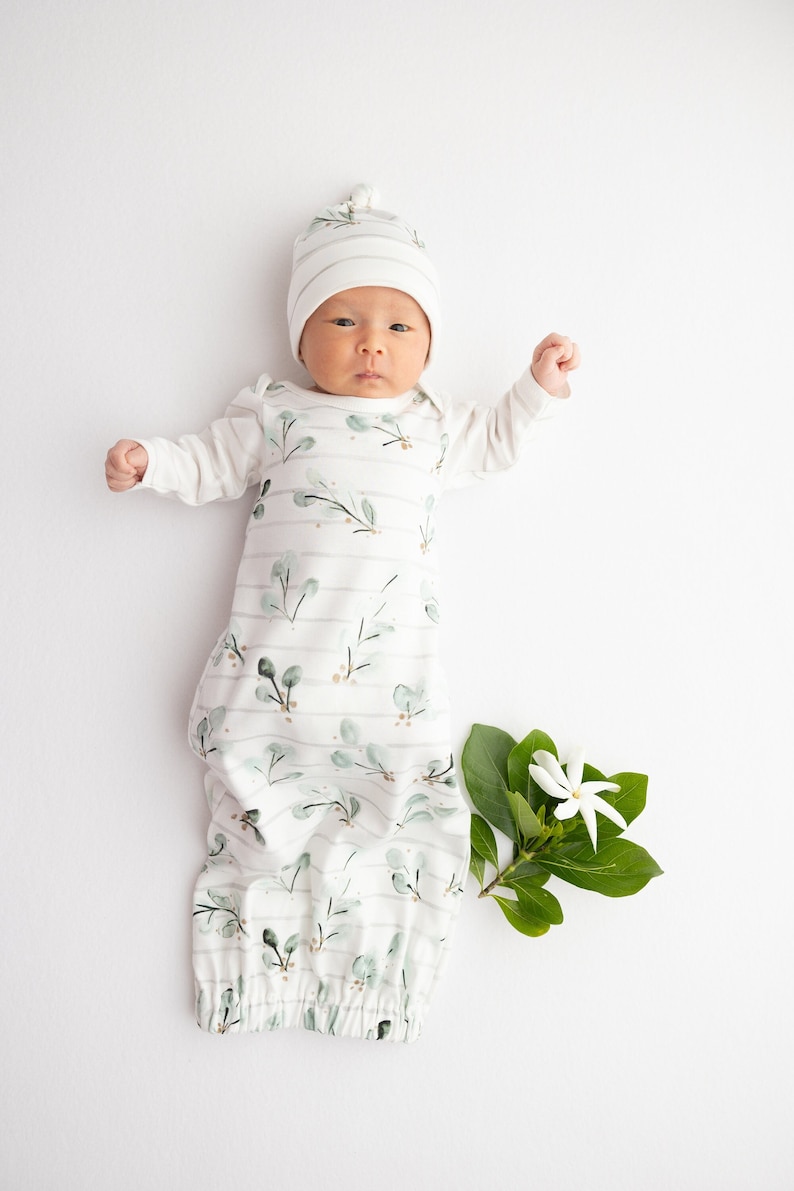 May include: A newborn baby wearing a white and green leaf-patterned sleep sack and matching striped hat. The baby is lying on a white surface with a sprig of leaves and a white flower.