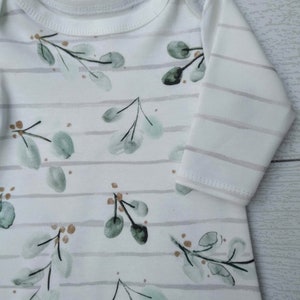 May include: White baby onesie with a botanical design. The onesie features a pattern of green eucalyptus leaves and small brown accents, set against a backdrop of horizontal gray stripes. The long-sleeved garment has a round neckline.