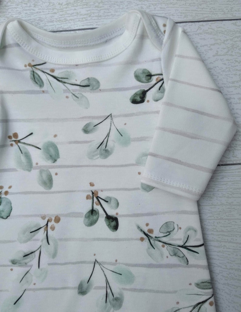 May include: White baby onesie with a botanical design. The onesie features a pattern of green eucalyptus leaves and small brown accents, set against a backdrop of horizontal gray stripes. The long-sleeved garment has a round neckline.
