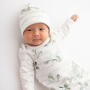 May include: A newborn baby wearing a white and gray striped onesie and matching hat. The onesie features a watercolor eucalyptus leaf design. The baby is lying on a white surface.
