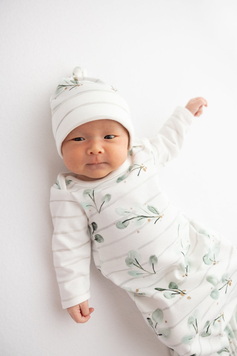 May include: A newborn baby wearing a white and gray striped onesie and matching hat. The onesie features a watercolor eucalyptus leaf design. The baby is lying on a white surface.