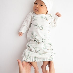 May include: A newborn baby wearing a white and green floral print sleep gown and matching striped hat. The gown has long sleeves and a ruffled hem. The baby is lying on a white surface.