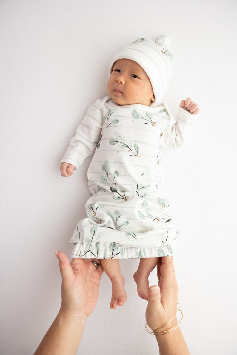 May include: A newborn baby wearing a white and green floral print sleep gown and matching striped hat. The gown has long sleeves and a ruffled hem. The baby is lying on a white surface.