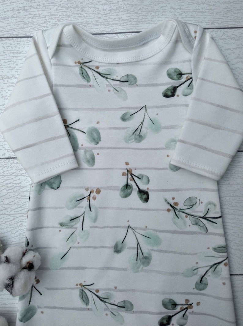 May include: White baby sleeper with long sleeves and a round neckline. The sleeper features a pattern of green eucalyptus leaves and brown accents on a white background with gray horizontal stripes.