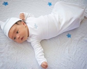 May include: A white baby gown with long sleeves and a pointed hat. The gown has the words "My Girl" embroidered on the front in blue thread. The baby is lying on a white sheet with blue star patterns.