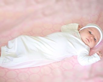 May include: A newborn baby wearing a white gown and a white knitted hat lies on a pink blanket with a circular pattern.