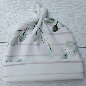 May include: A white baby knot hat with a watercolor eucalyptus leaf pattern in shades of green and gold. The hat features horizontal gray stripes and a folded brim. The top of the hat is tied in a knot.