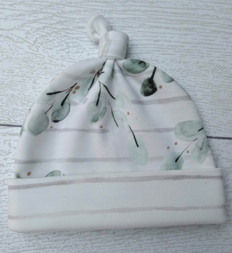 May include: A white baby knot hat with a watercolor eucalyptus leaf pattern in shades of green and gold. The hat features horizontal gray stripes and a folded brim. The top of the hat is tied in a knot.