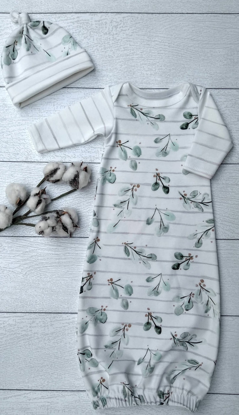 May include: A white baby gown and matching hat set. The gown and hat feature a watercolor-style botanical print with green leaves and brown accents, set against a striped background. The set is made of soft fabric.