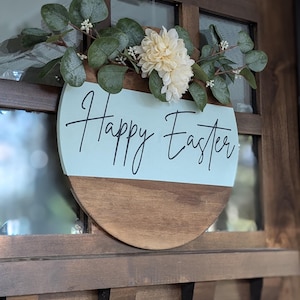 May include: A round wooden sign with a light blue background and the text "Happy Easter" in black script. The sign is decorated with a wreath of green leaves and white flowers.