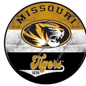 May include: A round, distressed-style graphic featuring the word "MISSOURI" in black letters at the top. The center displays a tiger head logo in black and gold. The word "Tigers" is written in gold script below the logo, with the year "1839".