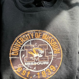 May include: Black sweatshirt with a brown and gold University of Missouri logo. The logo features a tiger head and the text "University of Missouri" and "Est. 1839".