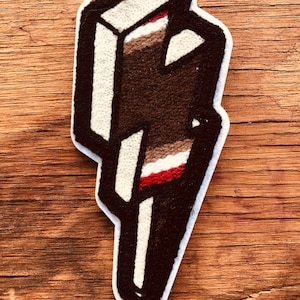 Puede incluir: Un parche bordado con forma de rayo, de color marrón, blanco y rojo. El parche está hecho de fieltro y tiene un contorno blanco.