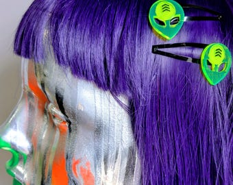 May include: Two neon green alien head hair clips attached to vibrant purple hair. Each clip features a black metal snap barrette base with a translucent, bright green acrylic alien face charm that includes etched chevron details on the forehead.