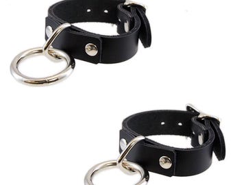 May include: Two black leather wrist cuffs with silver-colored metal rings and buckles. Each cuff has a wide band with a metal ring attached by a smaller metal loop. The cuffs are secured with a buckle and have silver-colored rivets.